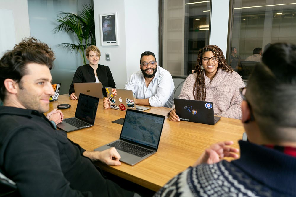 Six people sit around a conference table with laptops, smiling and engaged in a meeting in a modern office setting.