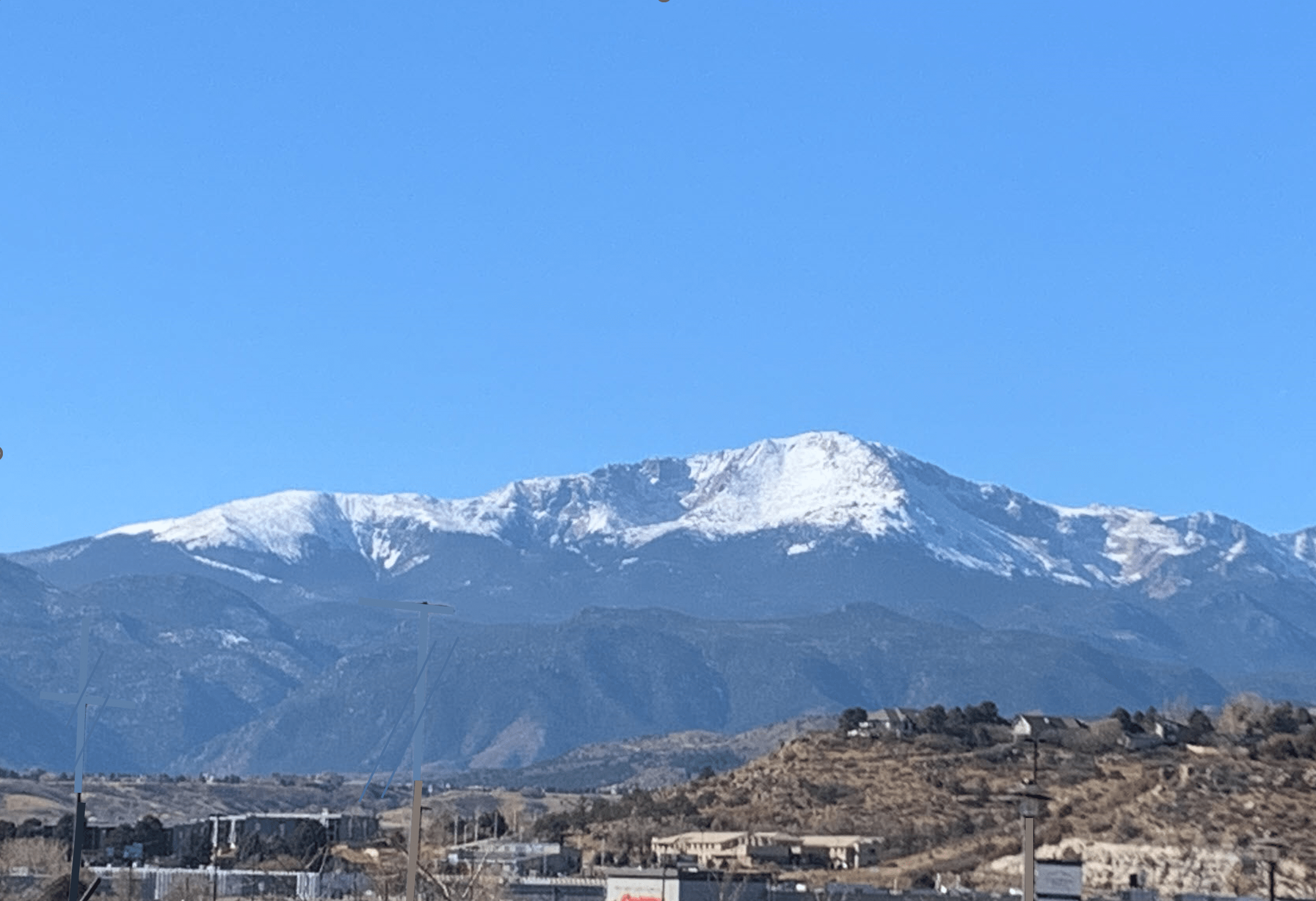 A scenic landscape featuring snow-capped mountains under a clear blue sky. There are scattered buildings and a few streetlights in the foreground, with green vegetation covering the hills at the base of the mountains.